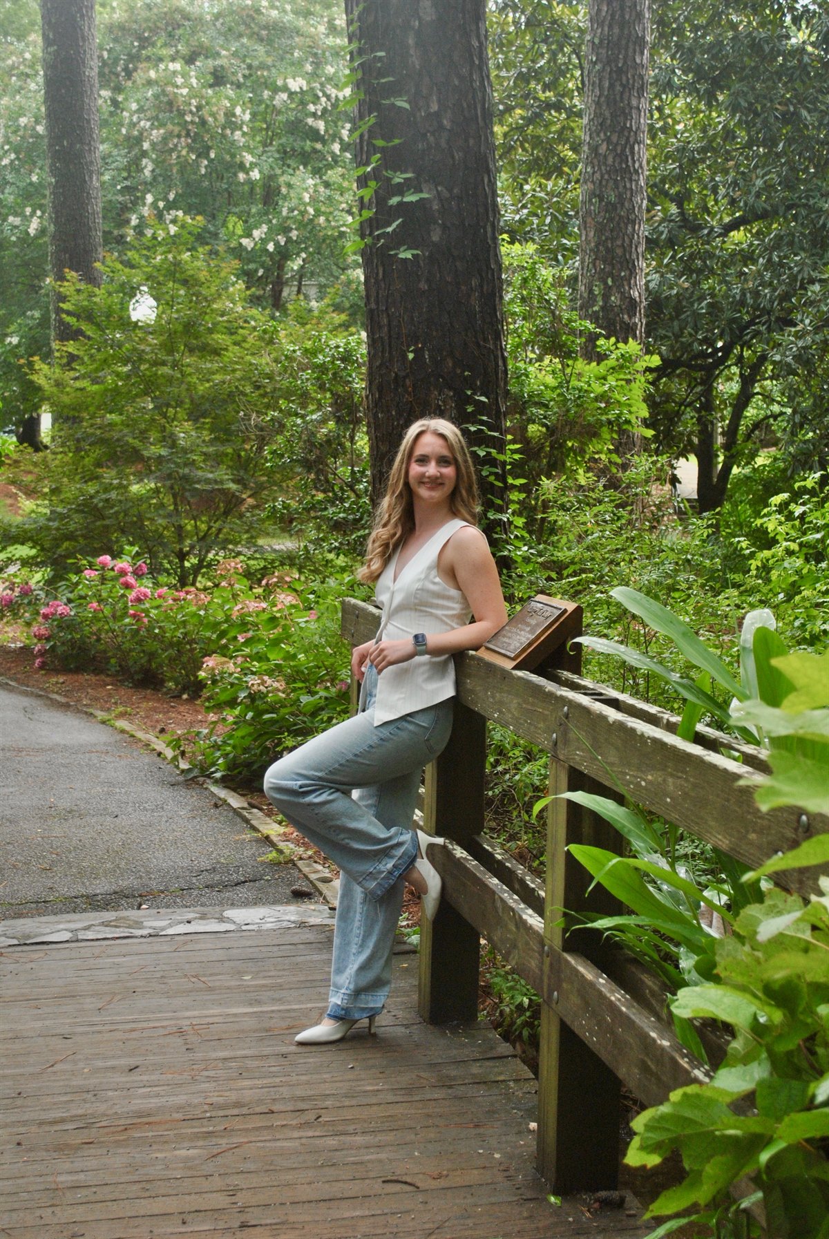 Piper Boss, founder of The BrightBrick Project, leaning against a wooden fence on a garden boardwalk surrounded by trees and flowers.